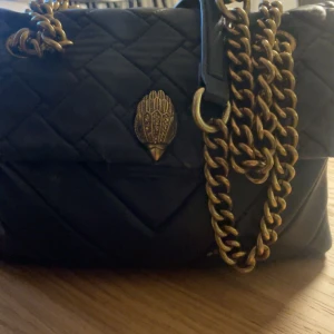 Svart mellanstor axelväska från Kurt Geiger - (Sliten på fodret, kolla bilderna)                       Säljer en snygg svart quiltad axelväska från Kurt Geiger, modell The Kensington. Väskan har guldfärgad kedja, dekorativt spänne framtill och är tillverkad i skinn. Perfekt storlek för det viktigaste och ger en cool vibe till din outfit. Denna har varit mycket använd, så den är rätt så sliten på insidan och på fodret på utsidan som man kan se på bilderna. Så den är sliten på utsidan och insidan, vilket man kan ha åtanke💞(skriv för fler bild