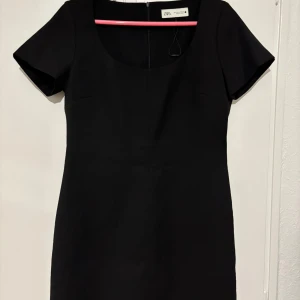 Svart kort klänning från Zara - En stilren svart kort klänning från Zara med rundad urringning och korta ärmar. Klänningen har en enkel, figurnära passform och dragkedja i ryggen. Perfekt för dig som gillar minimalistisk och klassisk stil. Kan användas med strumpbyxor