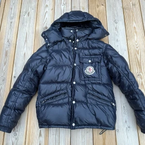 Svart Moncler pufferjacka med huva - Säljer en svart pufferjacka från Moncler med stor logga på bröstet, huva och dragkedjor på fickorna. Jackan har quiltad design, tryckknappar och en bröstficka med lock. Perfekt för kalla dagar och riktigt snygg streetstil.