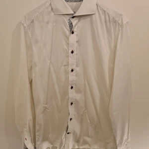 Vit skjorta Stenströms Slimline - Vit skjorta från Stenströms Slimline i bomull med snygga mörkblå knappar och röda knapphål. Insidan av krage och manschetter har ett stilrent blommigt mönster i blått. Skjortan har lång ärm och klassisk krage, perfekt för en clean och fräsch look.