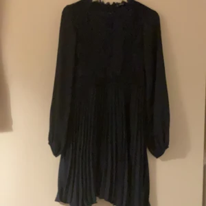 Mörkblå plisserad klänning med spets - Supersnygg mörkblå kortklänning med plisserad kjol och långärmade ärmar. Överdelen har fina spetsdetaljer och hög hals. Perfekt för dig som gillar en stilren look med extra detaljer. Kjolen är luftig och ger en cool rörelse. Oanvänd