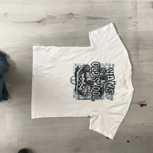 Vit Vans Old Skool t-shirt med tryck - Vit t-shirt från Vans i klassisk passform med stort grafiskt tryck på framsidan som visar ett par sneakers och texten 'Old Skool'. T-shirten har rund halsringning och korta ärmar. Materialet är mjuk bomull och trycket går i svart och blått.