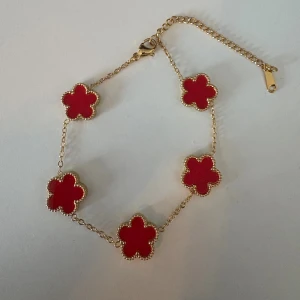 Guldarmband med röda blommor - Superfint armband med fem röda blommor i klöverform, infattade i guldfärgad kedja. Blommorna har en snygg pärlad kant och armbandet har justerbar längd med karbinlås. Perfekt för dig som vill sticka ut med färg och cool design.