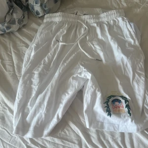 Vita shorts från Casablanca med tryck - Säljer ett par vita shorts från Casablanca med elastisk midja och snörning. På ena benet finns ett färgglatt tryck med logga och motiv. Sköna och luftiga, perfekta för varma dagar. Materialet känns mjukt och lätt.