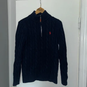 Mörkblå stickad tröja Polo Ralph Lauren - Mörkblå kabelstickad tröja från Polo Ralph Lauren med halv dragkedja och hög krage. Tröjan har lång ärm, ribbade muddar och röd broderad logga på bröstet. Perfekt för kyliga dagar och snygg till jeans eller chinos.