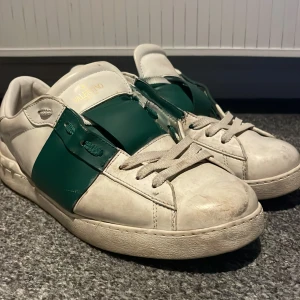 Valentino Garavani sneakers vit/grön - Snygga Valentino Garavani sneakers i vitt skinn med bred grön rem över mitten och coola nitar på hälen. Klassisk rund tå, platt sula och vita snören. Perfekta för dig som vill sticka ut med en lyxig touch och streetkänsla. Tvättar före lämning de är F@  ke pris kan diskuteras