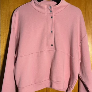 Rosa sweatshirt med knappar - Säljer en ljusrosa sweatshirt med hög krage och snygga tryckknappar framtill. Tröjan har långa ärmar och en relaxed, loose passform. Perfekt för dig som gillar en chill och trendig stil.