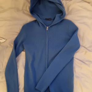 Blå ribbad zipper med luva från Brandy Melville - Säljer en blå ribbstickad hoodie från Brandy Melville med dragkedja hela vägen och huva. Tröjan har långa ärmar och är gjord i mjukt stickat material. Perfekt för dig som gillar en chill och snygg look. Med luva