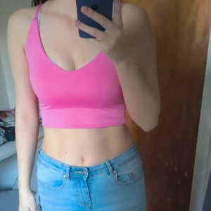 Rosa croppad linnetopp - Säljer en croppad rosa linnetopp med smala axelband och djup v-ringning. Toppen har en tight passform som framhäver midjan. Perfekt för sommardagar eller när du vill ha en färgklick i din outfit. Har en bit mitt i v-ringningen som är sprucken men går att laga. Sen en nopra back på ryggen.