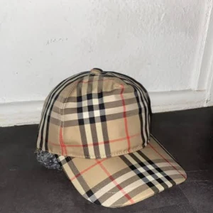 Burberry keps - Snygg keps från Burberry i klassiskt rutigt mönster med beige, svart, vitt och röda linjer. Justerbar baksida med tryckknappar och Burberry London England-patch. Perfekt accessoar för att lyfta din outfit. Priset kan diskuteras 
