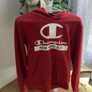 Röd hoodie från Champion - Röd hoodie från Champion med stor vit logga och texten 'New York N.Y.' framtill. Klassisk känguruficka och huva. Perfekt för dig som gillar streetwear och vill ha en snygg och bekväm tröja med sportig vibe. I fint använt skick