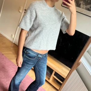 Ljusgrå stickad tröja från Gina Tricot - Supermjuk ljusgrå stickad tröja från Gina Tricot med kort ärm och boxig, croppad passform. Perfekt till jeans eller kjol och har en enkel, stilren look med ribbade kanter vid hals och ärmslut.