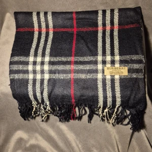 Rutig ullhalsduk från Burberry - Klassisk rutig halsduk från Burberry i marinblått, vitt och röda detaljer. Gjord i 100% lammull och har fransar i kanterna. Den ikoniska Burberry-taggen sitter synligt på ena sidan. Perfekt accessoar för att lyfta din outfit.