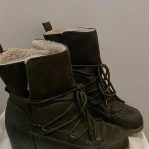 Olivgröna moon boots med foder ”inuiiki”  - Säljer ett par olivgröna moon boots med mjukt beige foder och snörning runt hela skon. Utsidan är i mocka och skinn, med en platt sula och rund tå. Perfekta för kalla dagar när du vill hålla stilen och värmen.