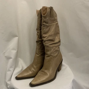 Beiga cowboy boots i skinn - Snygga beiga cowboy boots i mjukt skinn med spetsig tå och markerad klack. Klassiska broderade detaljer och rynkad skaftdesign ger en cool western-vibe. Perfekta för dig som vill sticka ut med en trendig och avslappnad look. Boho-chic