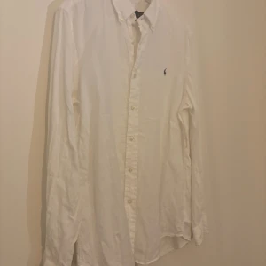 Vit skjorta från Polo Ralph Lauren - Klassisk vit skjorta från Polo Ralph Lauren med button down-krage och broderad logga på bröstet. Skjortan har lång ärm, knappar framtill och är tillverkad i linne. Perfekt för en clean och stilren look.