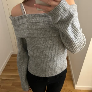 Grå offshoulder stickad tröja från H&M - Snygg grå stickad tröja med ribbad struktur och offshoulder-design. Tröjan har långa ärmar och bred kant upptill som ger en cool och avslappnad vibe. 