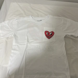 Vit Keith Haring t-shirt med hjärta - Vit t-shirt från Keith Haring med ett stort rött hjärta med ansikte tryckt på bröstet och en mindre version på fickan. Klassisk passform med korta ärmar och rund hals. Tillverkad i mjuk bomull, perfekt för dig som gillar streetart och coola prints.