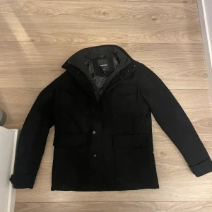 Svart parkajacka  - Svart parkajacka med hög krage och flera stora fickor framtill. Jackan har lång ärm, knappar och dragkedja samt ett quiltat innerfoder för extra värme. Perfekt för kalla dagar och har en clean, stilren look.