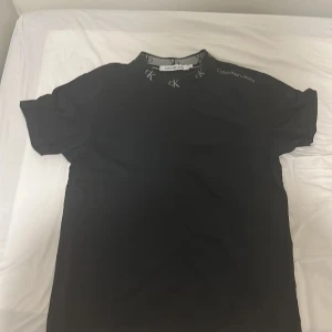 Svart t-shirt Calvin Klein Jeans XS - Svart t-shirt från Calvin Klein Jeans i bomull. Klassisk modell med korta ärmar och rund halsringning med CK-logga runt kragen. Diskret Calvin Klein Jeans-tryck på axeln. Perfekt för en clean och stilren look.