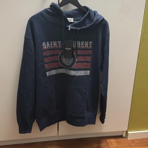 Mörkblå Saint Laurent hoodie - Säljer en mörkblå hoodie från Saint Laurent med tryckt logga och ränder i rött och vitt på bröstet. Tröjan har huva med snörning och en stor magficka. Perfekt för dig som gillar streetstyle och vill ha något snyggt och bekvämt.