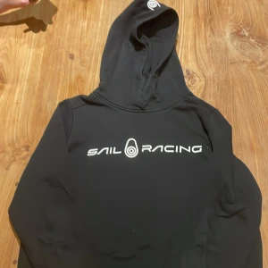 Svart Sail Racing hoodie med tryck - Svart hoodie från Sail Racing med stort vitt logotryck på bröstet. Klassisk modell med huva och långa ärmar. Perfekt för dig som gillar sportig och clean stil. Materialet känns mjukt och följsamt mot kroppen. Pris kan diskuteras