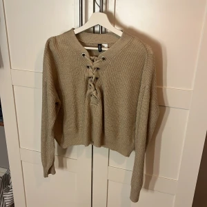 Beige stickad tröja med snörning H&M - Beige stickad tröja från H&M Divided med snygg snörning framtill och grovstickad struktur. Modellen är croppad med långärmade ärmar och bred ribbad mudd vid ärmslut och nederkant. Perfekt för lager-på-lager under hösten.