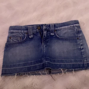 Blå jeanskjol med broderi, stl 36 - Säljer en blå jeanskjol i kort modell även low waist med rå kant nertill och snygga slitningar. Kjolen har klassiska fickor fram och bak, samt ett broderat mönster med pärlor på ena bakfickan. Har använt 1 gång. 