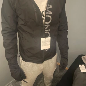 Svart vindjacka från Moncler med huva - Svart vindjacka från Moncler med stor logga längs framsidan och klassisk Moncler-patch på ärmen. Jackan har huva, dragkedja framtill och två sidofickor med dragkedja. Tillverkad i polyester och har en clean, sportig look.