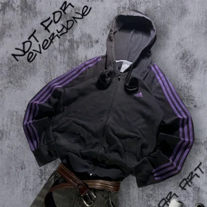 Svart Adidas hoodie med lila ränder - Svart hoodiejacka från Adidas med lila klassiska ränder längs ärmarna och Adidas-logga på bröstet. Jackan har huva med dragsko, dragkedja framtill och två fickor. Perfekt för en sportig och avslappnad stil.