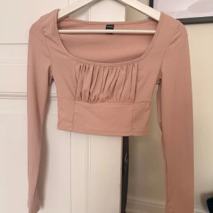 Ribbad croppad topp  - Säljer en ljusrosa ribbad croppad topp med långa ärmar och snygg rynkdetalj framtill. Toppen har en fyrkantig ringning och är figurnära i modellen, perfekt att styla med höga jeans eller kjol. Den har en liiiten liten fläck längst ner till höger på sista bilden. Sänker därav priset. 