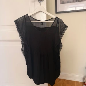 Svart blus med volang  - Svart blus från H&M Mama med transparenta partier upptill och längs ärmarna. Blusen har volangdetaljer längs axlarna och är ärmlös med rund halsringning. Luftigt och lätt material som ger en snygg och bekväm look.