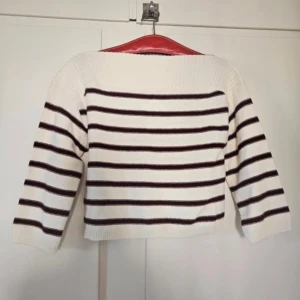 Randig stickad tröja från H&M Divided - Vit stickad tröja med mörkröda horisontella ränder från H&M Divided. Tröjan har en bred ribbad halsringning och trekvartsärmar. Perfekt för dig som gillar en chill och stilren look med lite fransk vibe.