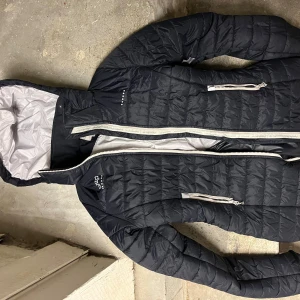 Svart lång pufferjacka från Uhip - Säljer en mörkblå pufferjacka från Uhip med huva och vita detaljer. Jackan har flera dragkedjor, quiltad design och är lång i modellen. Perfekt för kalla dagar och har en snygg, sportig look med praktiska fickor och reflekterande detaljer.