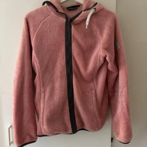 Rosa fleecejacka från OnThePeak - Mysig rosa teddyjacka från OnThePeak med huva. . Jackan har hel dragkedja framtill, två sidofickor och svarta detaljer vid dragkedjan och huvan. Perfekt för kyliga höst och vår dagar.  Hund och katt finns i hemmet 