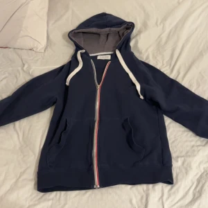 Mörkblå hoodie med dragkedja från Pier One - Säljer en mörkblå hoodie från Pier One med vit dragsko och hel dragkedja framtill. Tröjan har två fickor och en huva med grått foder. Perfekt för en avslappnad stil.