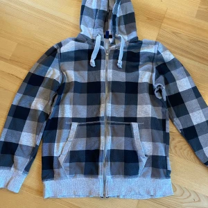 Rutig hoodie från H&M DIVIDED - Säljer en rutig hoodie från H&M DIVIDED i svart och grått. Tröjan har dragkedja, huva med snörning och två fickor framtill. 