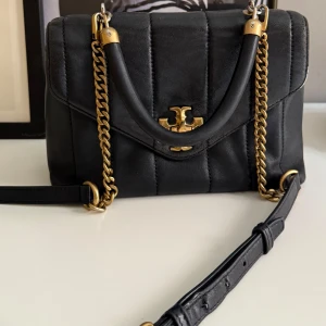 Tory Burch Kira small - Snygg svart handväska från Tory Burch i quiltat läder med guldfärgade kedjedetaljer och logotyp framtill. Väskan har både handtag och axelrem, samt praktiska fack och dragkedja inuti. Perfekt storlek för det viktigaste och riktigt stilren design.
