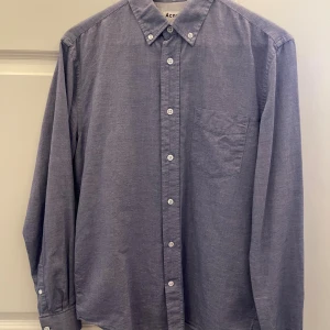 Blå skjorta från Acne Studios - Snygg blå skjorta från Acne Studios med klassisk button-down krage och vita knappar. Skjortan har en bröstficka och är tillverkad i mjuk bomull. Passformen är normal och ärmarna är långa. Perfekt för dig som gillar stilrena och enkla plagg.