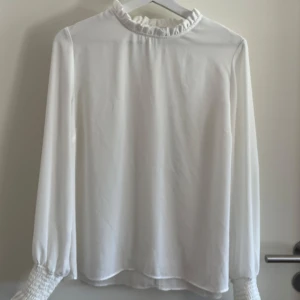 Vit blus från Gina Tricot  - Vit blus i storlek 40 från Gina Tricot. Volanger längs halsen som är högre än normalt och volanger på ärmmändarna. 