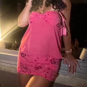 Rosa glittrig kort kjol och topp - Superfin rosa kort 2 delad  med glittriga detaljer och blommigt mönster. Axelbandslös modell med söt rosettliknande twist vid bysten och figurnära passform upptill. Perfekt för dig som vill sticka ut och glänsa.