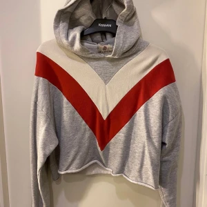 Grå croppad hoodie från Pull&Bear EU M - Grå croppad hoodie från Pull&Bear med stor huva och V-format mönster i rött och vitt över bröstet. Tröjan har långa ärmar och avslappnad passform, perfekt för en chill look. Materialet är mjuk bomullsmix och den har ribbade muddar vid ärmsluten.