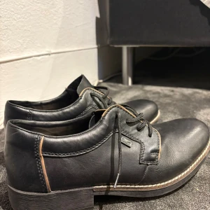 Svarta skinnskor från Rieker, storlek 42 - Klassiska svarta skinnskor från Rieker med snörning och robust sula. Skorna har diskreta kontrastsömmar och en låg klack, perfekt för en stilren look. Märket RiekerTX syns på sidan och sulan är rejält mönstrad för bra grepp.