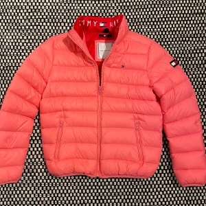 Rosa dunjacka Tommy Hilfiger - Lätt rosa dunjacka från Tommy Hilfiger med vattenavvisande yta. Dragkedja framtill och två sidofickor med dragkedja. Insidan är röd med logotyp och innerficka. Enkel att packas ner i ryggfickan. Mycket bra skick!