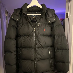 Svart pufferjacka från Polo Ralph Lauren - Svart pufferjacka från Polo Ralph Lauren i storlek XS (xl barn) men passar exakt som en xs. Jackan har huva, flera dragkedjor och ett rött Polo-logo broderat på bröstet. Bra jacka nu för de kalla dagarna, behåller värmen enormt bra. Inga defekter eller skador som jag hittat, tveka inte att höra av er vid funderingar eller frågor! 
