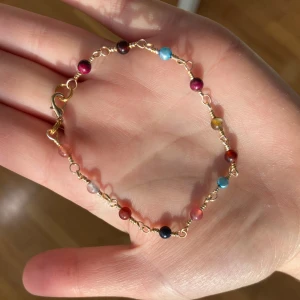 Färgglatt pärlarmband med guldlänk - Snyggt armband med små runda pärlor i blått, rött, svart och rosa på tunn guldfärgad kedja. Perfekt för dig som gillar färg och vill ha något unikt på handleden. Justerbar storlek med flera öglor för låset. Stenarna är turmalin. 