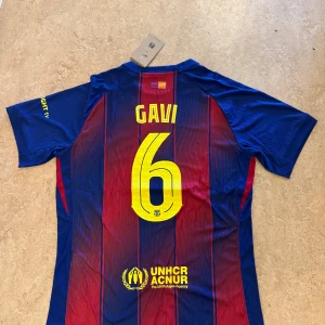 FC Barcelona Gavi matchtröja Nike L - Säljer en FC Barcelona matchtröja med Gavi #6 på ryggen. Tröjan är kortärmad, tillverkad i Dri-Fit material och har klassiska blå och röda vertikala ränder. Framsidan har klubbmärke, Nike-logga och Spotify-logga i gult. Perfekt för dig som älskar Barca!