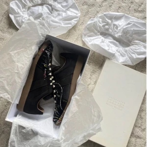  Maison Margiela sneakers med färgstänk - Svarta sneakers i skinn med unikt färgstänkt mönster i flera färger. Skorna har beige snörning och en klassisk gummisula. Perfekt för dig som vill sticka ut med en kreativ och modern stil.