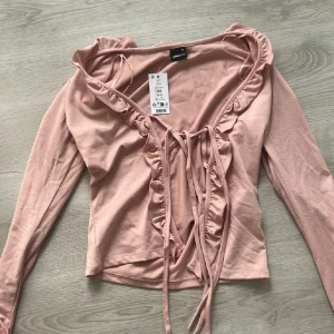 Ljusrosa volangtop från Gina Tricot - Supersöt ljusrosa bolerotopp från Gina Tricot med volangdetaljer längs halsringningen och ärmsluten. Toppen har långa ärmar, croppad passform och knyt framtill. Perfekt för dig som gillar romantiska och trendiga plagg.