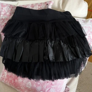 Svart volangkjol i satin och tyll - Supersnygg svart kortkjol med flera lager volanger i både satin och tyll. Kjolen har bred midja och ger en cool, fluffig look med mycket volym. Perfekt för dig som vill sticka ut med en edgy och trendig stil.